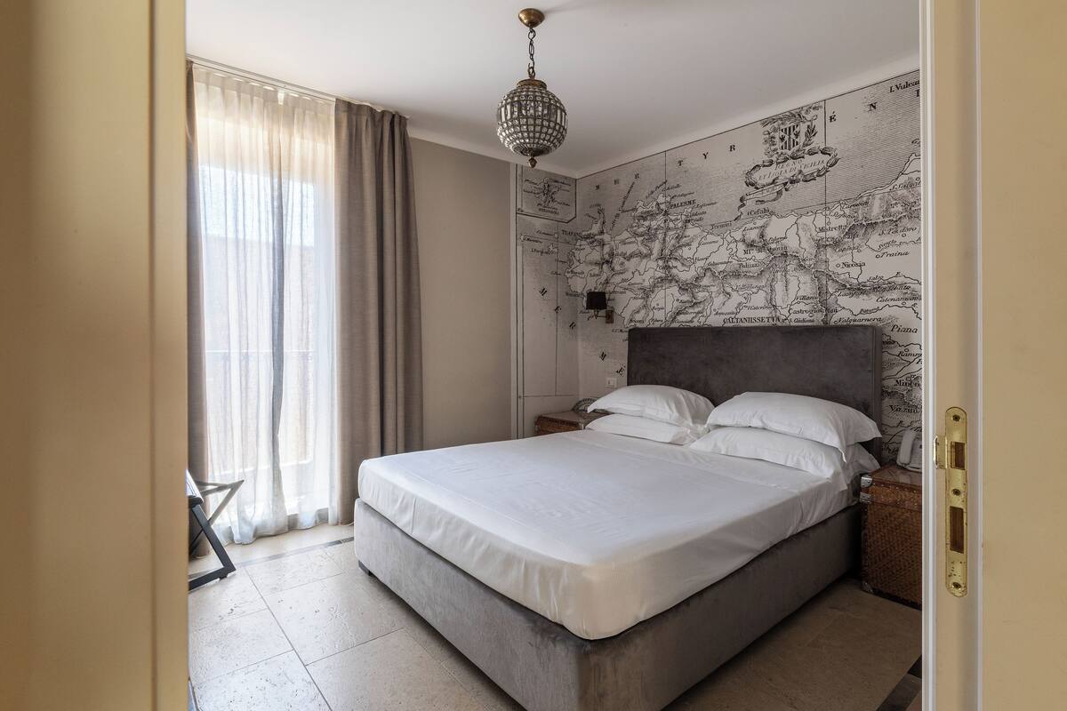 A neatly made bed in a room with a large map on the wall, a chandelier, and a view of the outside through sheer curtains.