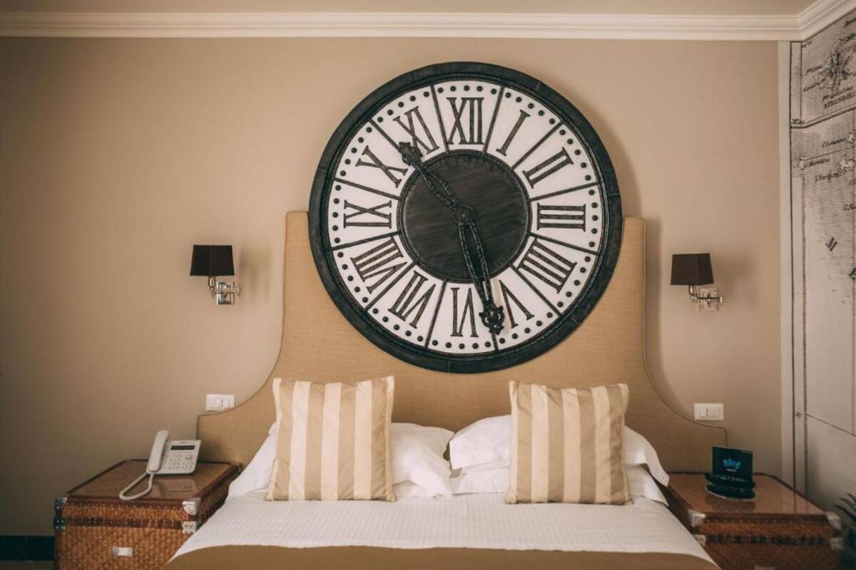 A hotel room with a large wall clock, a bed with striped pillows, a bedside telephone, and a small table with a lamp.
