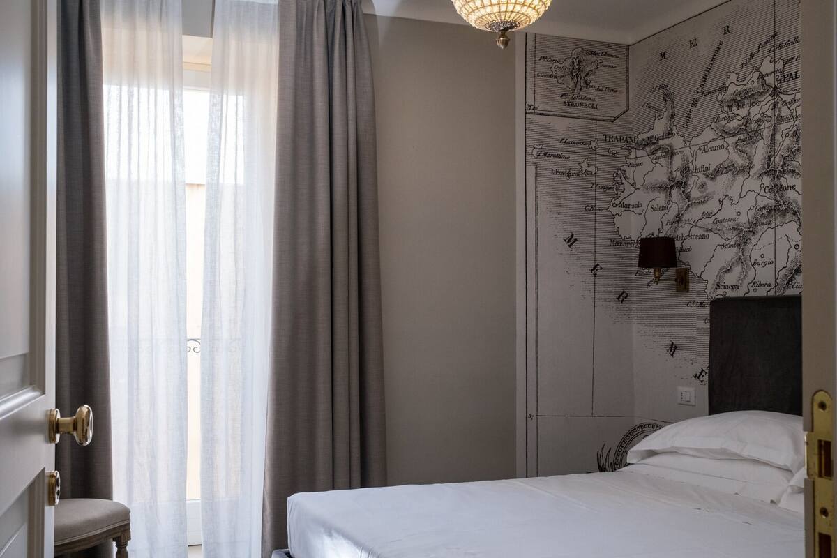 A hotel room with a bed, a map on the wall, a window with curtains, and a chandelier.