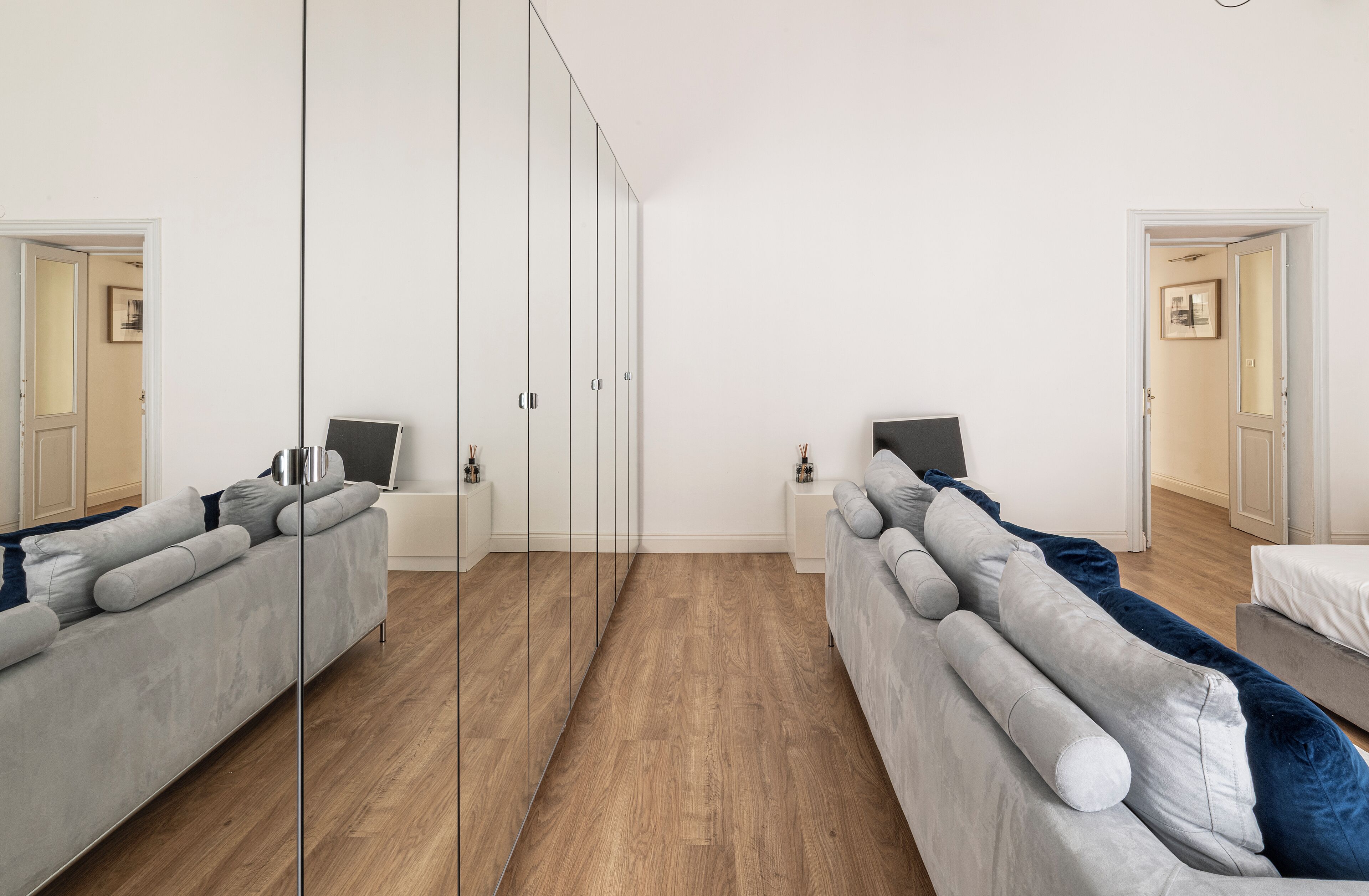 A modern, minimalist bedroom with a large mirror, two grey sofas, a wooden floor, and a white wardrobe.