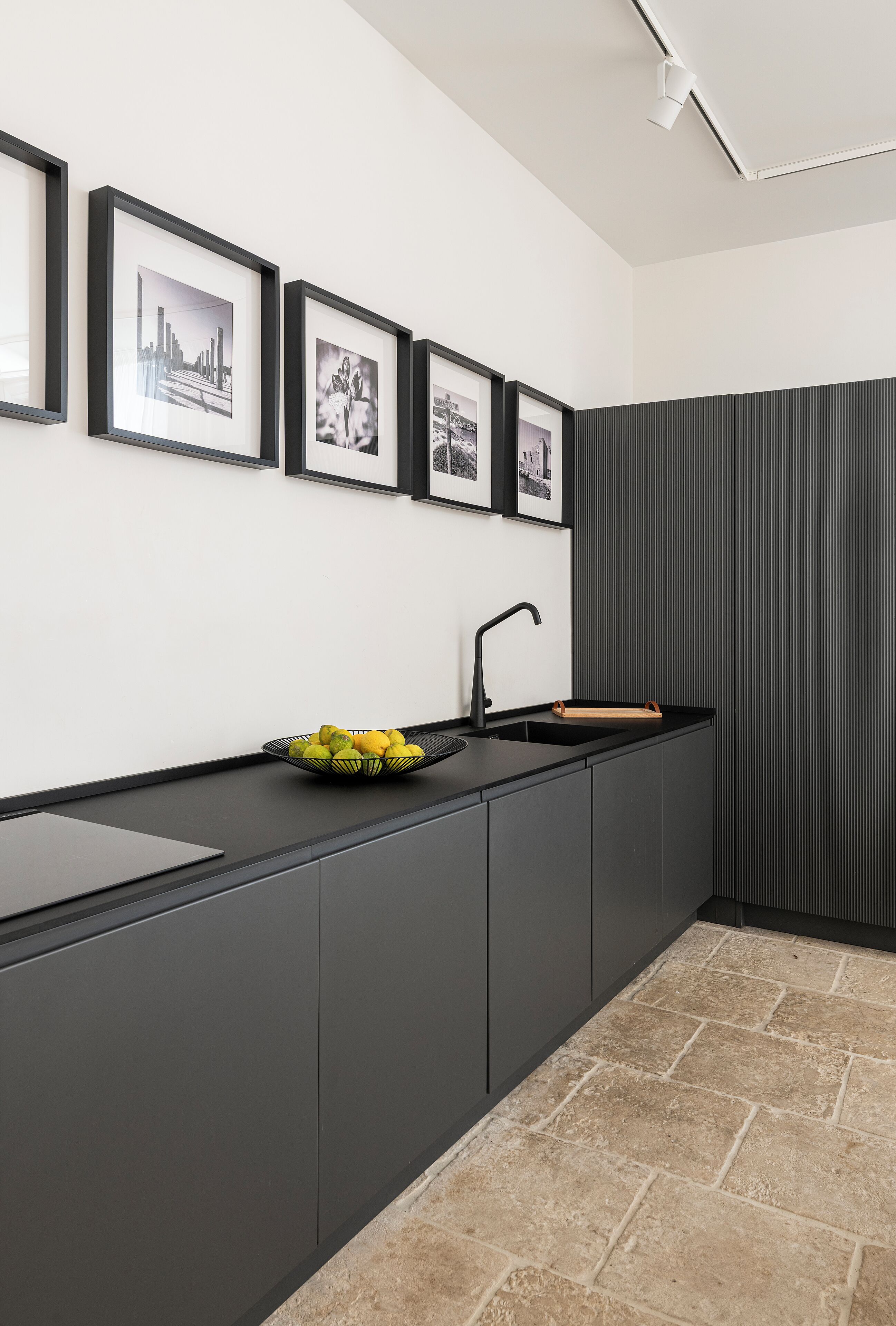 A modern kitchen with a black countertop, a sink, and a bowl of fruit. There are framed pictures on the wall above the counter.