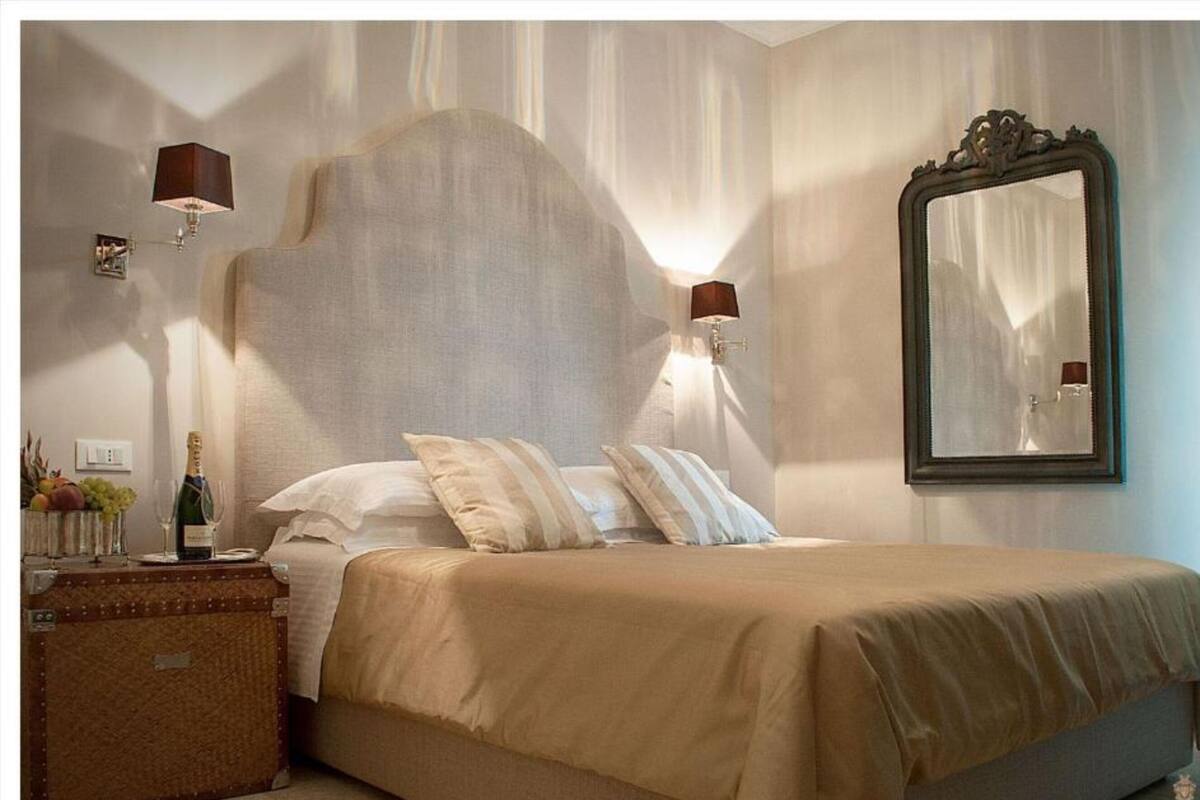 A bedroom with a large bed, two wall-mounted lamps, a mirror, and a bedside table with a bottle and glasses.