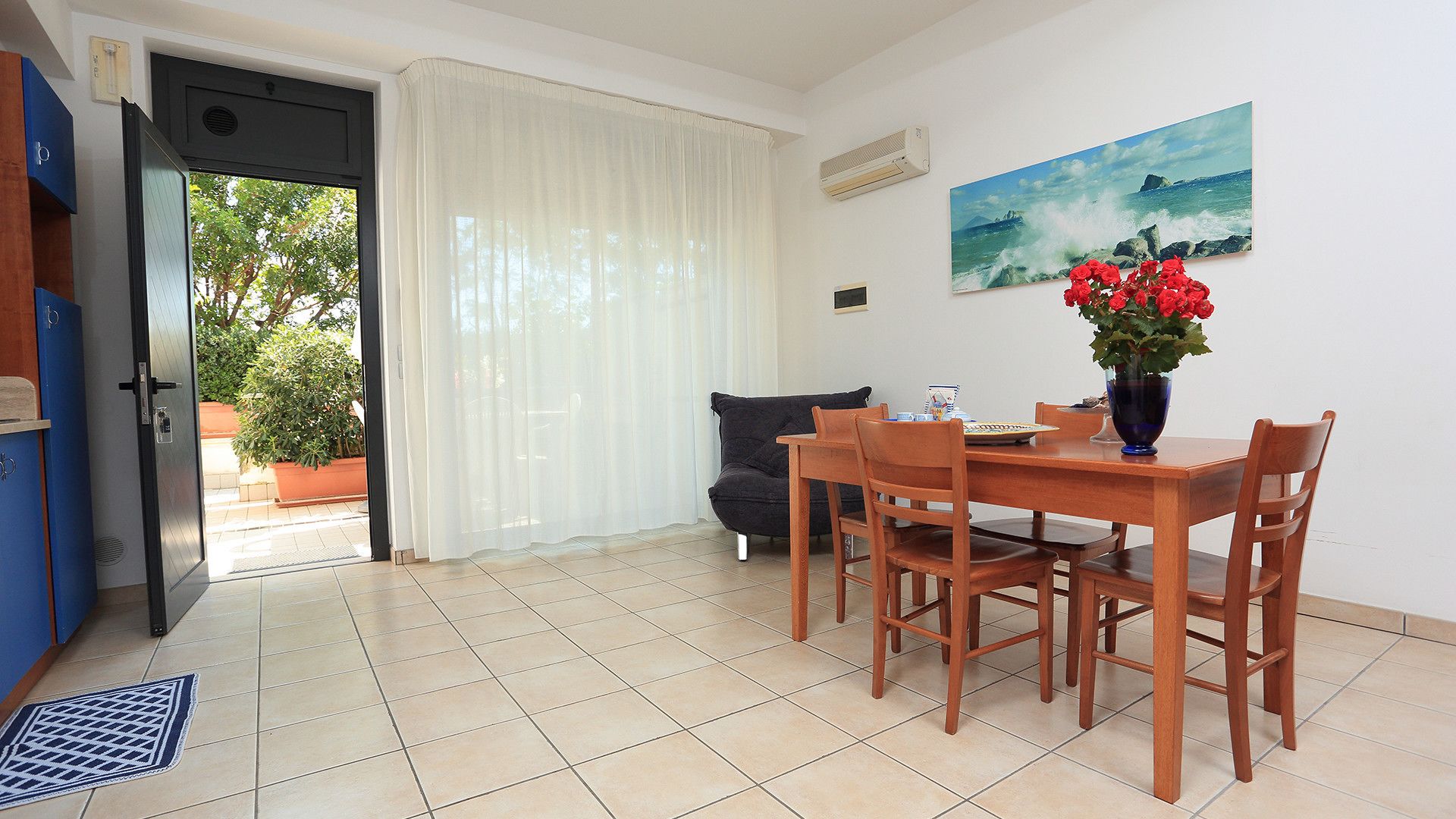 A dining area with a table and chairs, a sofa, a painting, and a door leading to an outdoor area with plants.