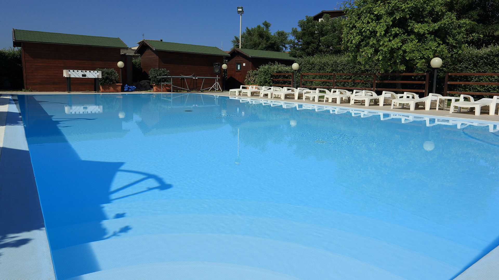 A clear swimming pool with white lounge chairs around it, wooden cabins in the background, and a clear blue sky.