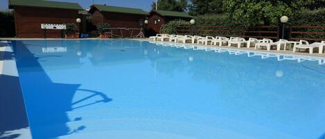 A clear swimming pool with white lounge chairs around it, wooden cabins in the background, and a clear blue sky.
