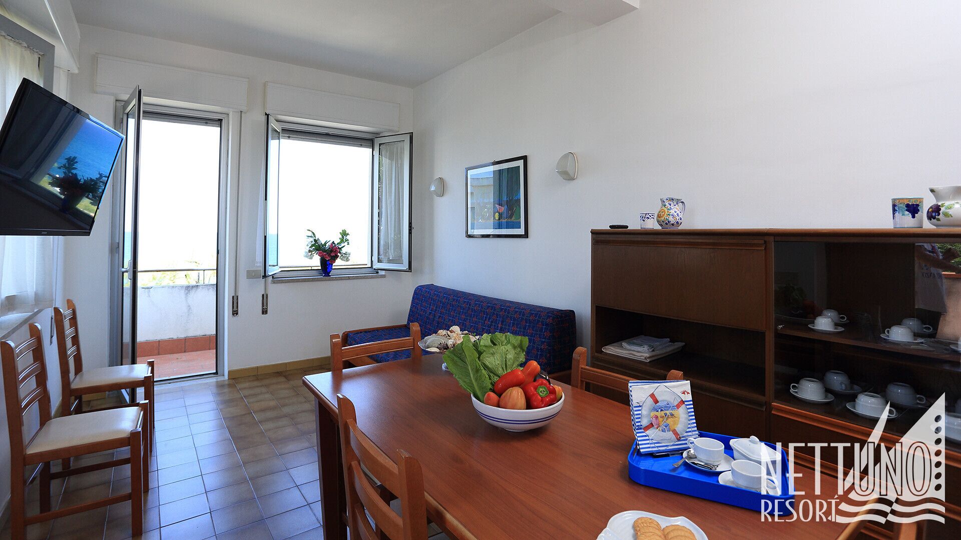 A dining area with a wooden table, chairs, a bowl of fruit, a blue tray with cups, and a bookshelf with tea sets.