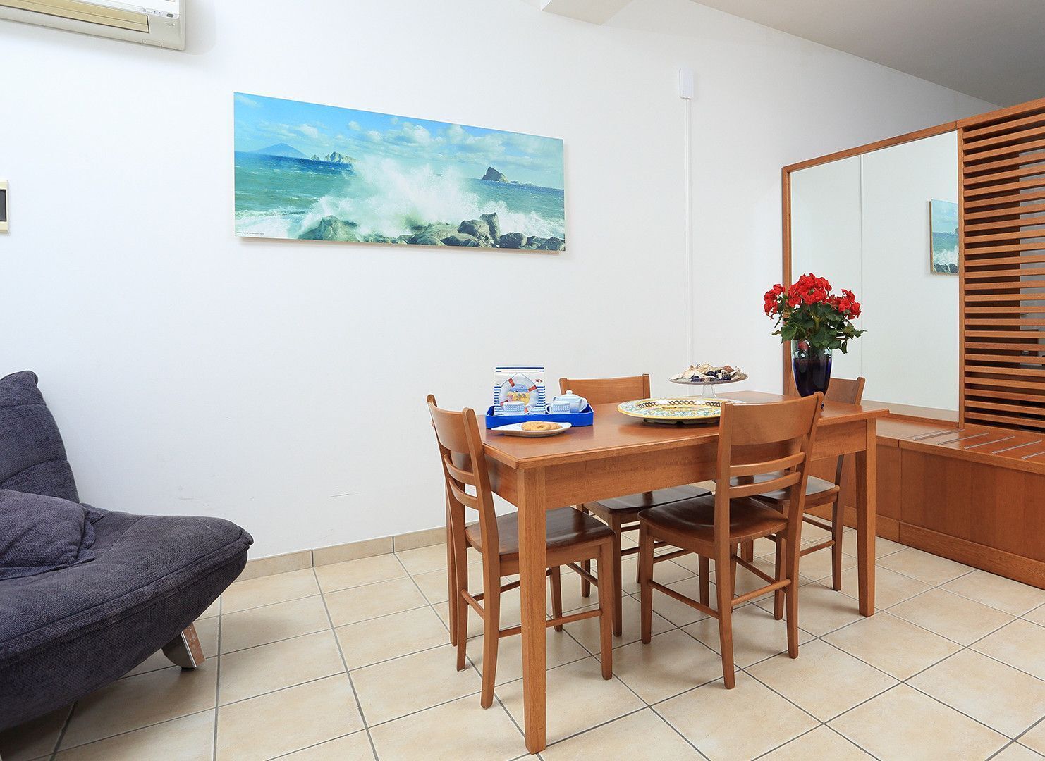 A dining area with a wooden table and chairs, a painting of a coastal scene, and a vase with red flowers.