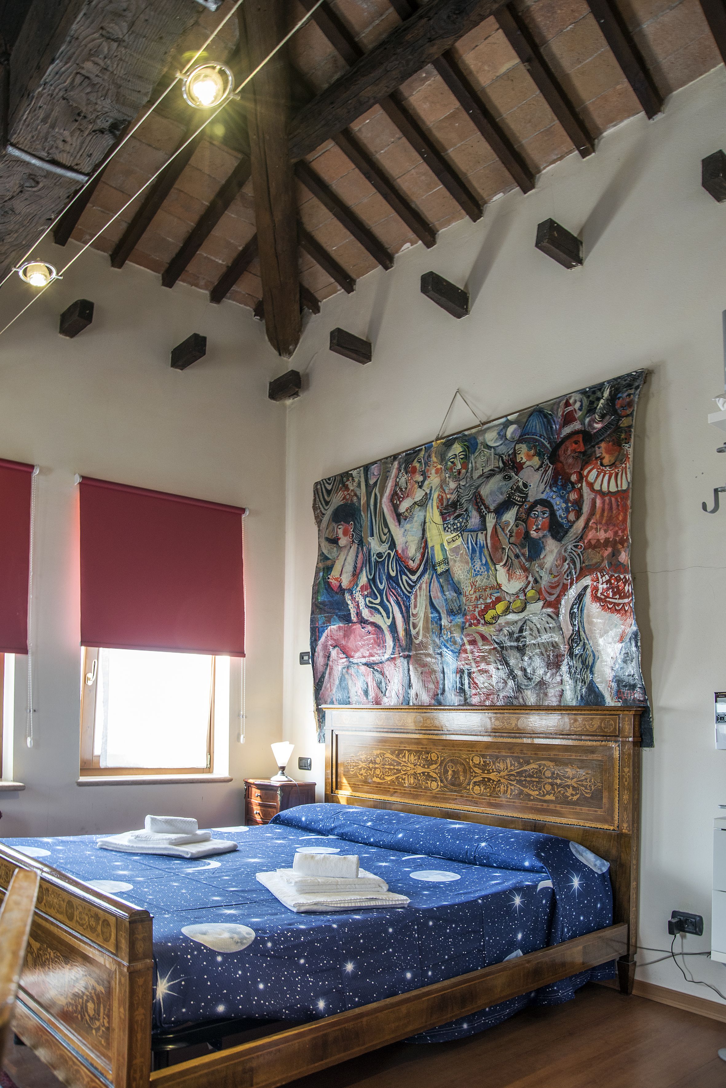 A bedroom with a wooden bed, a large tapestry on the wall, and a window with red blinds.