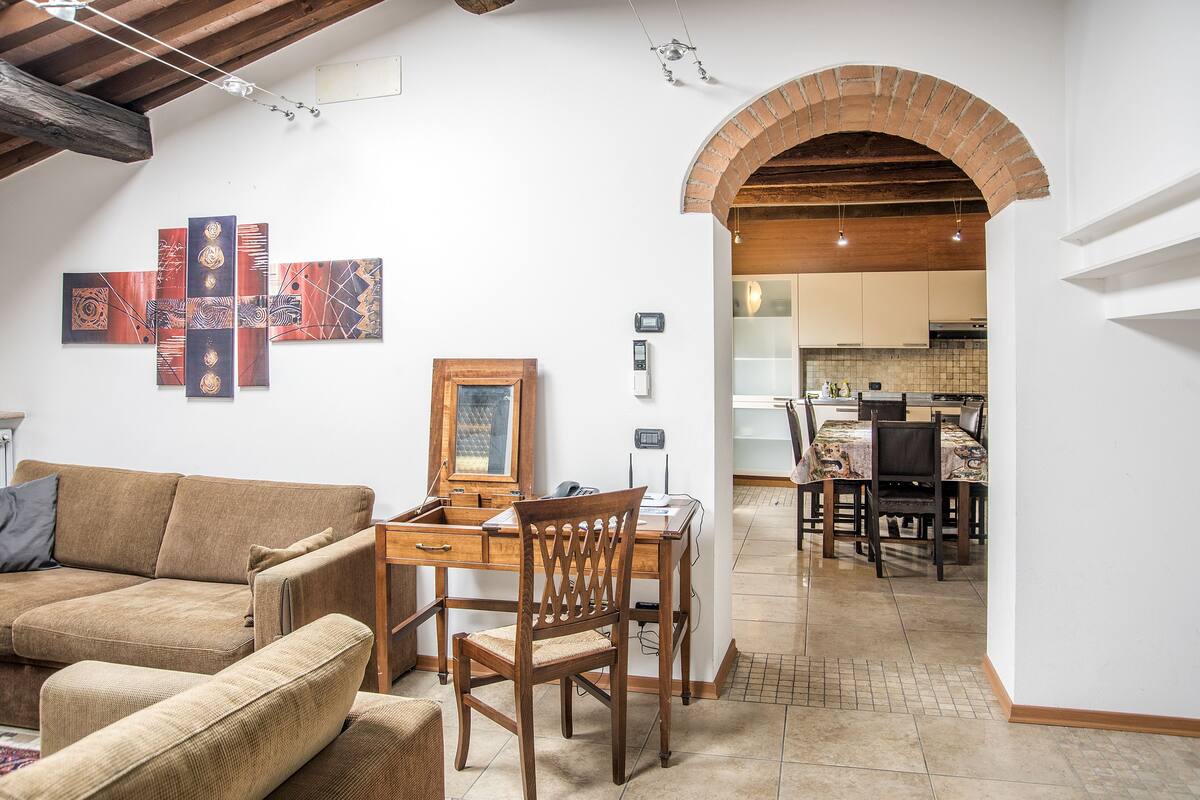 A cozy living room with a wooden dining table, a wooden chair, and a sofa. There is a kitchen visible through an archway.
