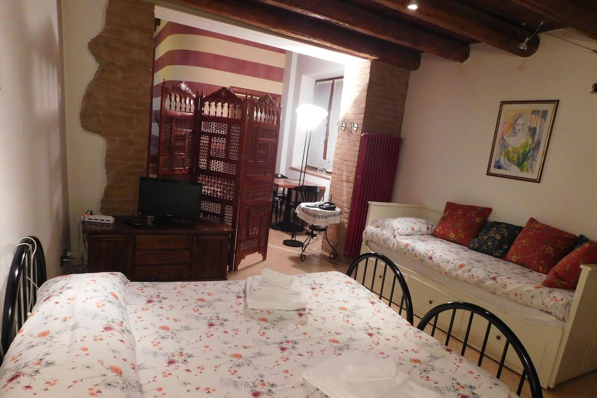 A bedroom with a bed, a desk with a TV, a small table with a chair, and a sofa.