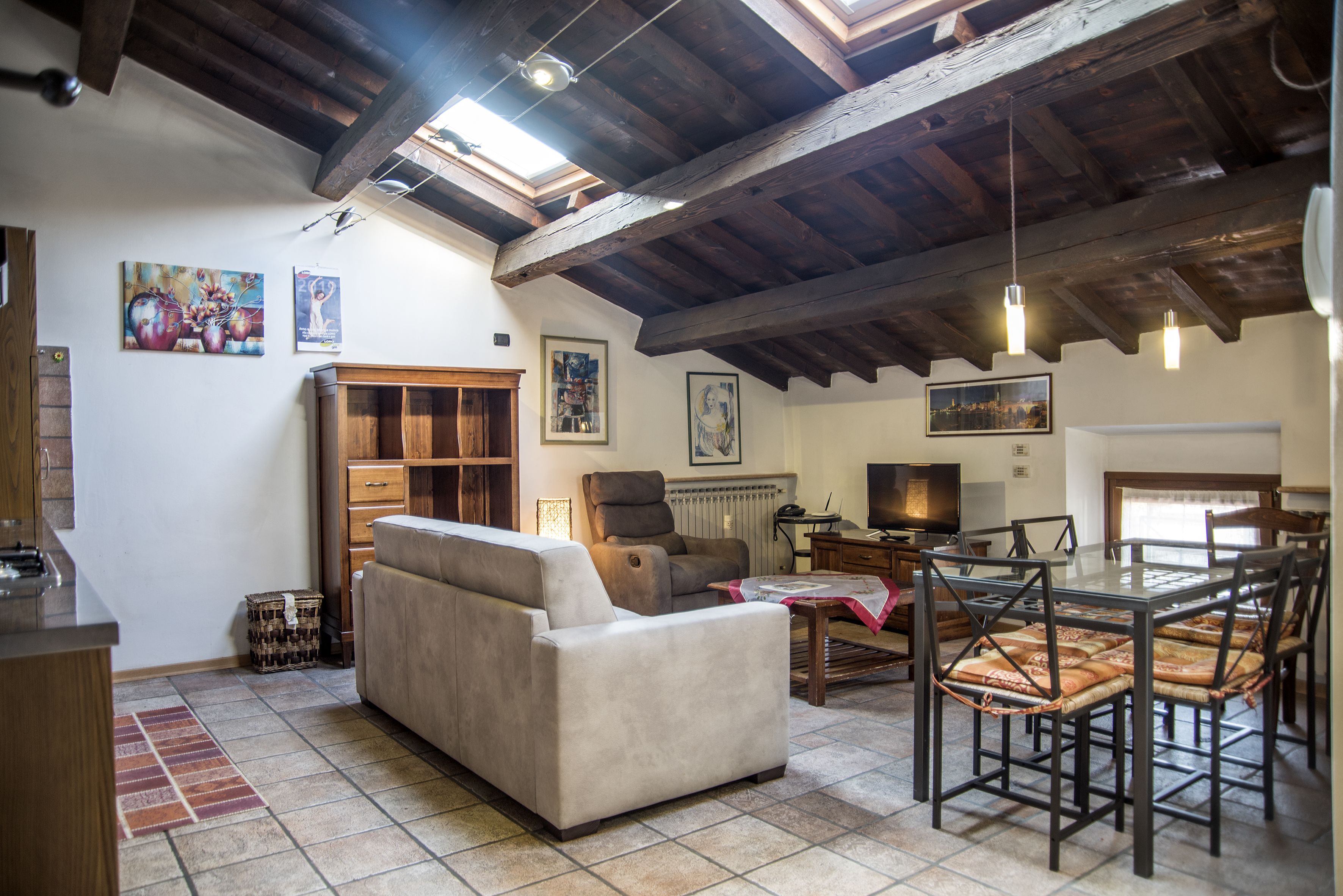 A spacious living area with a sofa, dining table, and chairs, wooden beams on the ceiling, and artwork on the walls.