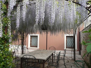 Un espacio de estar al aire libre cubierto con una mesa y sillas, rodeado por una pérgola cubierta de glicinas.