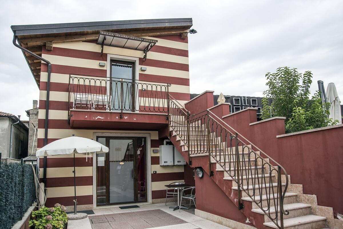 A two-story building with a red and white striped facade, a balcony with metal railings, a staircase with a metal railing, and a small outdoor seating area with a table and chairs.