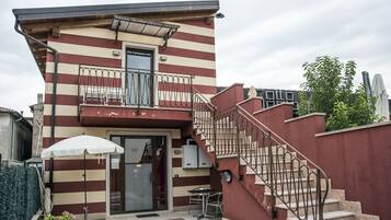 Un bâtiment à deux étages doté d’une façade à rayures rouges et blanches, d’un balcon avec des rampes en métal, d’un escalier avec une rampe en métal et d’un petit espace extérieur aménagé avec une table et des chaises.