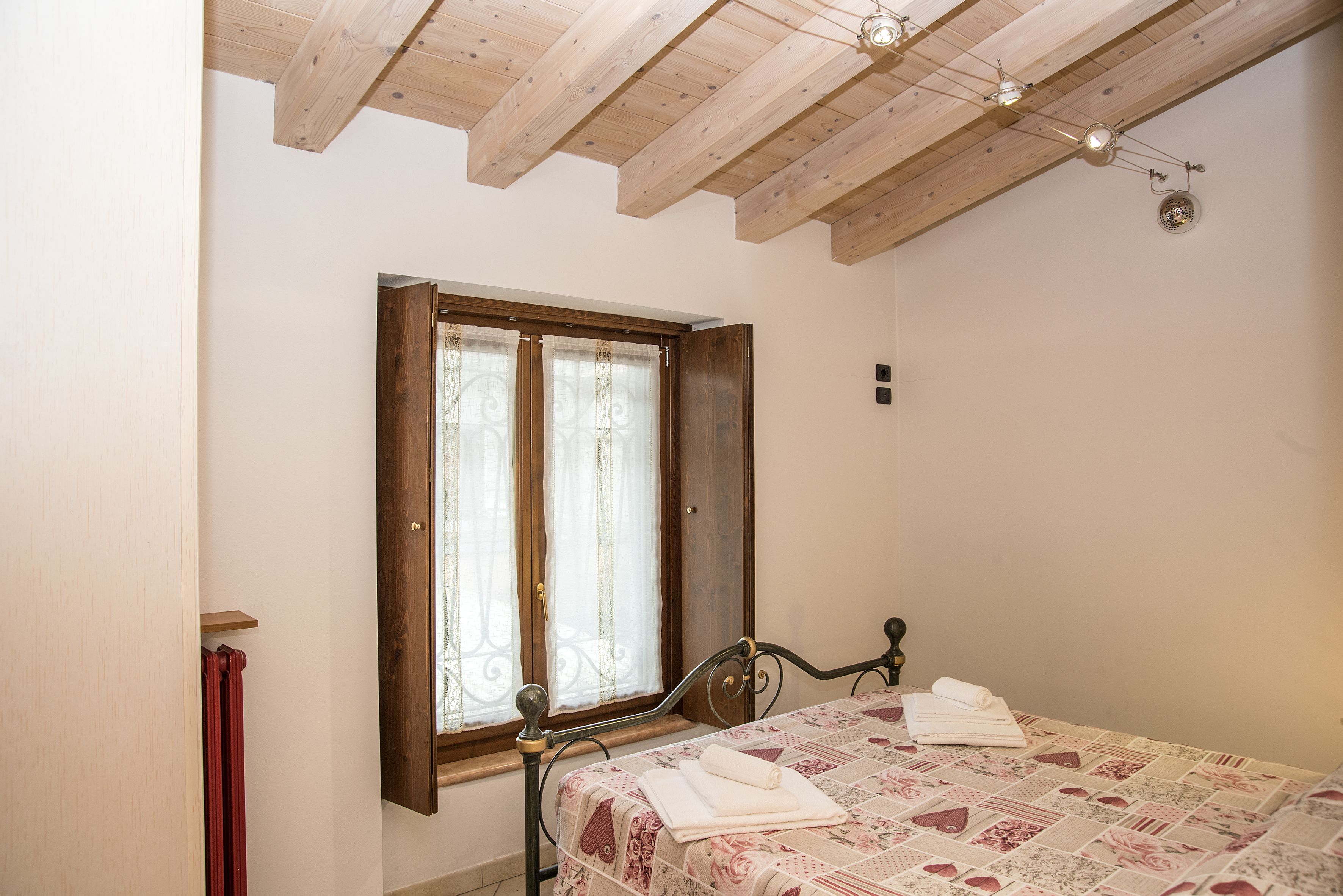 Quarto com teto de madeira, cama com colcha florida, janela com cortinas e radiador.