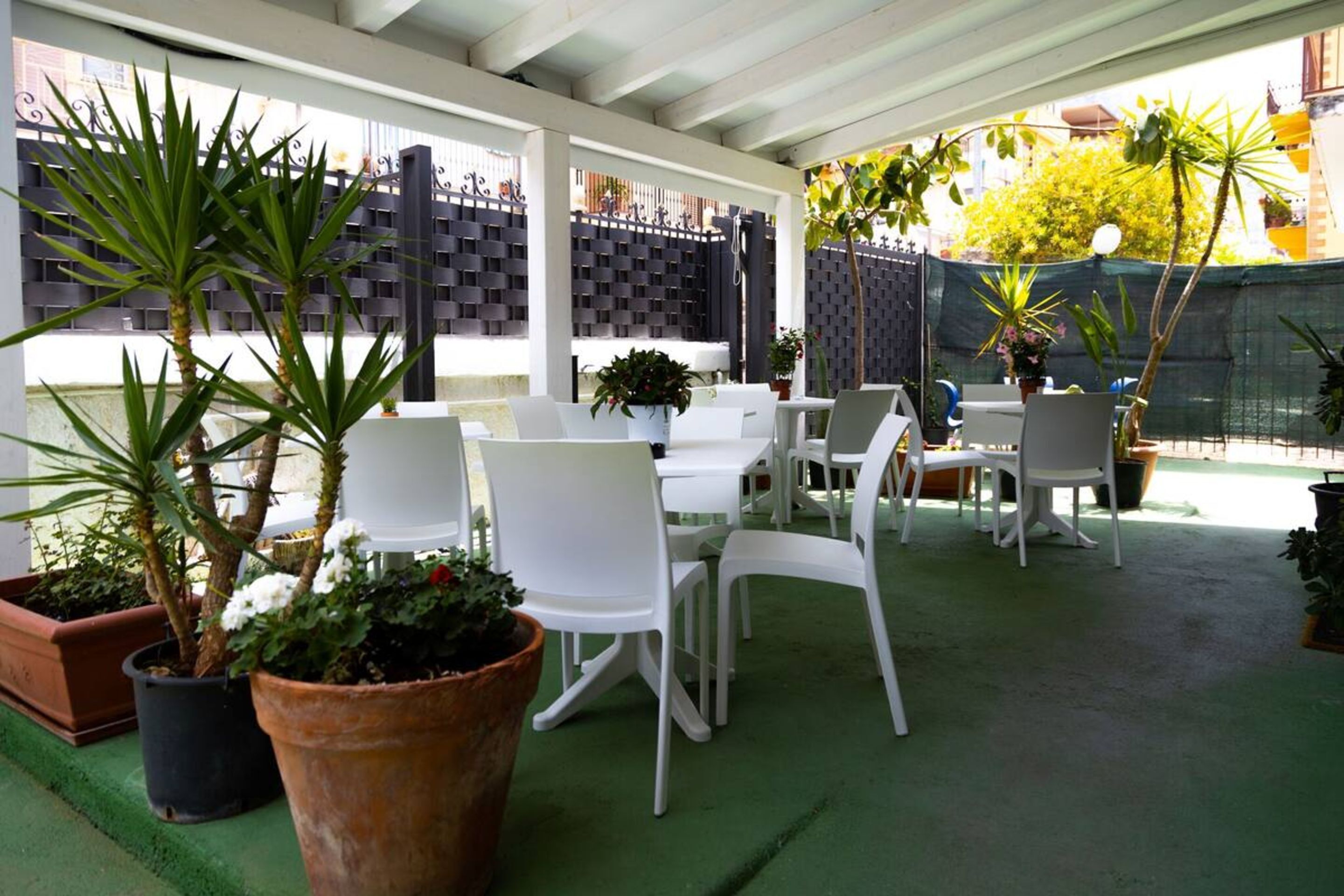 An outdoor patio area with white plastic chairs and tables, surrounded by potted plants and a green artificial turf floor.