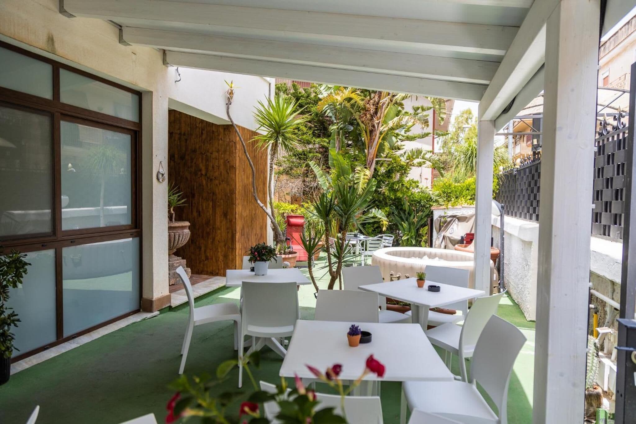 Un patio al aire libre con mesas y sillas blancas, rodeado de plantas en macetas y una valla de madera.