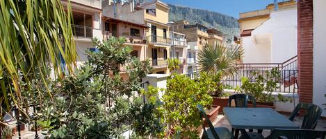 A terrace with a table and chairs, surrounded by potted plants and overlooking a residential area with buildings and a mountain in the background.