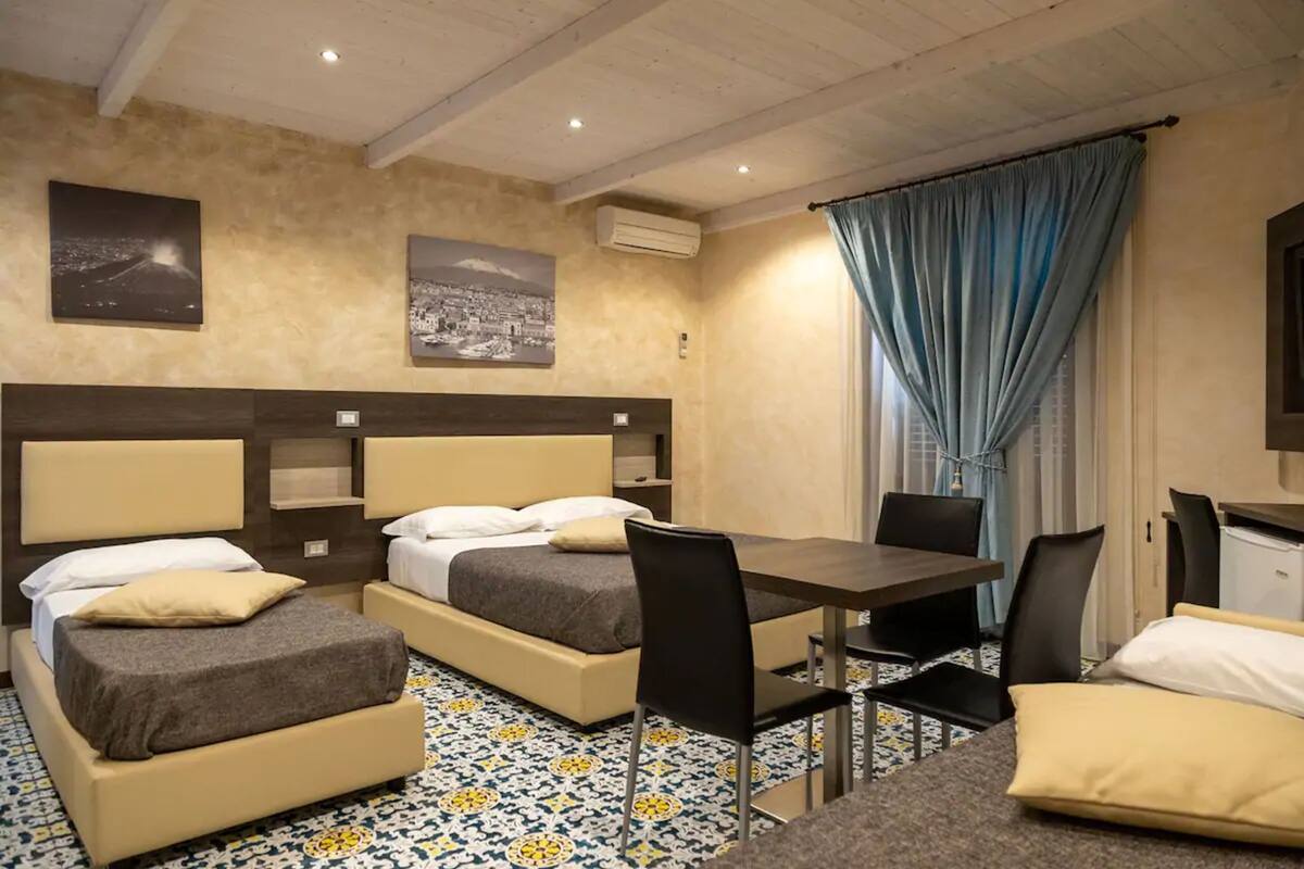 A hotel room with two beds, a dining table, chairs, a small kitchenette, and artwork on the walls.