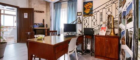 Un comedor con una mesa de madera, sillas y un cuenco de fruta. Hay un escritorio con monitor, teclado y una campanilla de bronce. El cuarto tiene piso de madera, una escalera con barandilla decorativa y una pared con cuadros enmarcados.