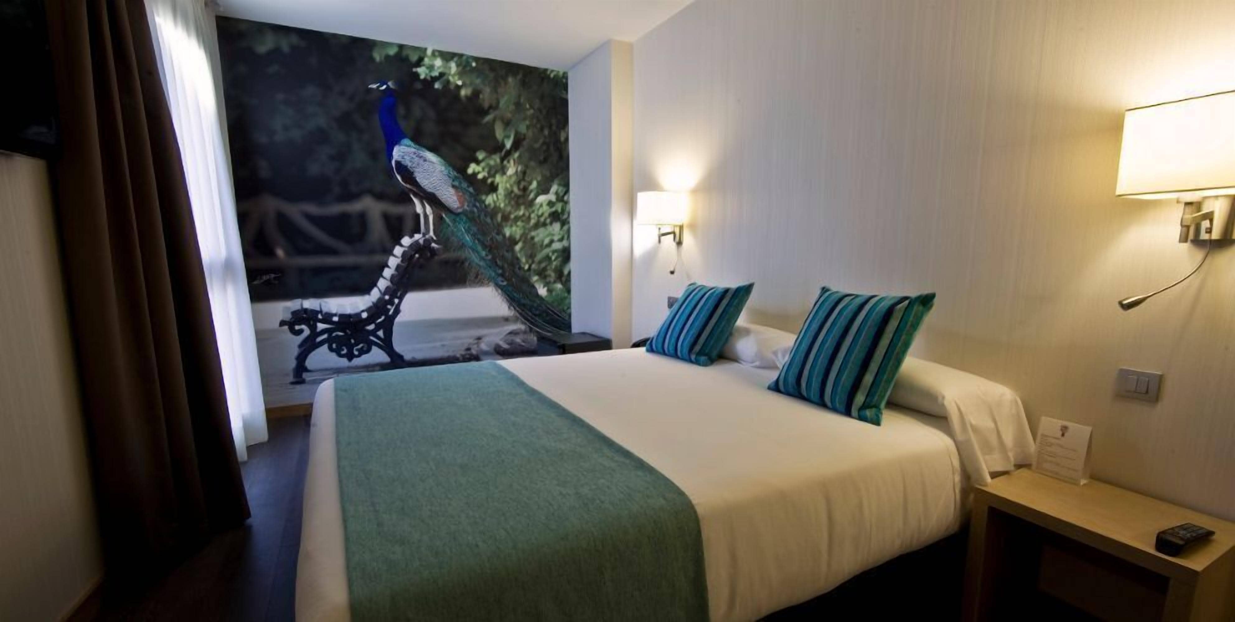 Habitación de hotel con una cama grande, una mesita de noche, una lámpara de pared y un mural de pavo real.