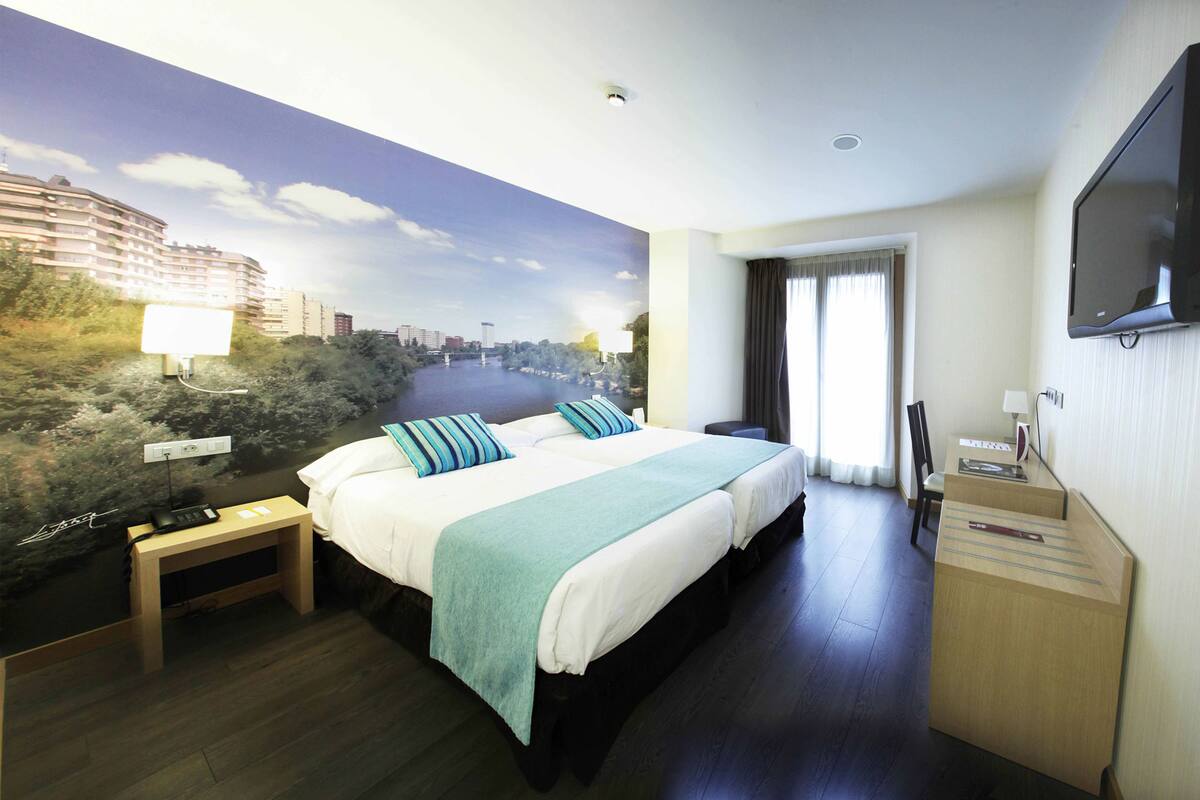 A hotel room with a large bed, a desk, a flat-screen TV, and a mural of a cityscape.
