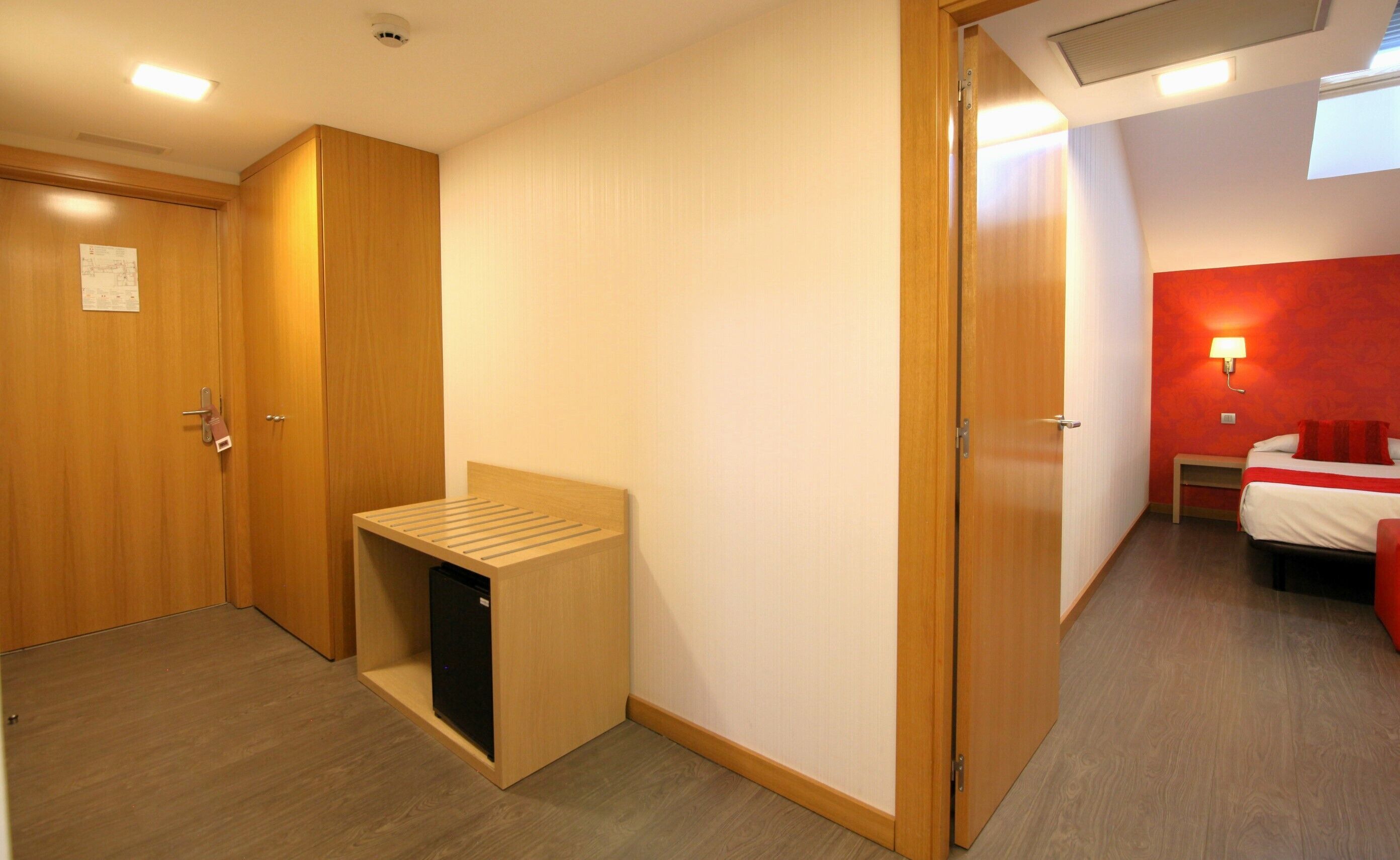 A hotel room with a wooden wardrobe, a small bench, a compact refrigerator, a bed with red bedding, and a nightstand.