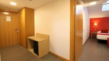 A hotel room with a wooden wardrobe, a small bench, a compact refrigerator, a bed with red bedding, and a nightstand.