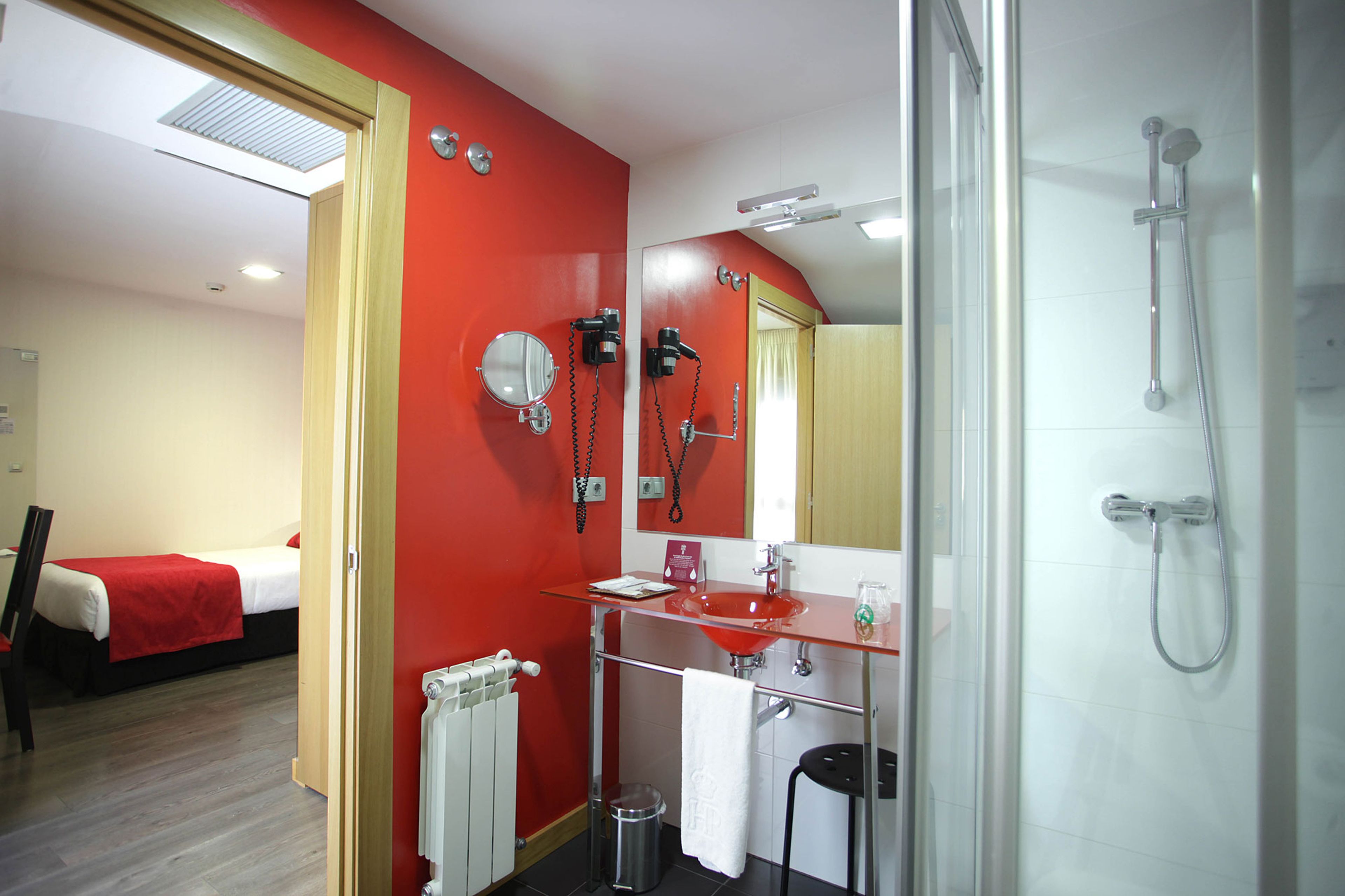 Une salle de bain d’hôtel avec un mur rouge, un lavabo en verre, un miroir, une douche et un lit dans la pièce adjacente.