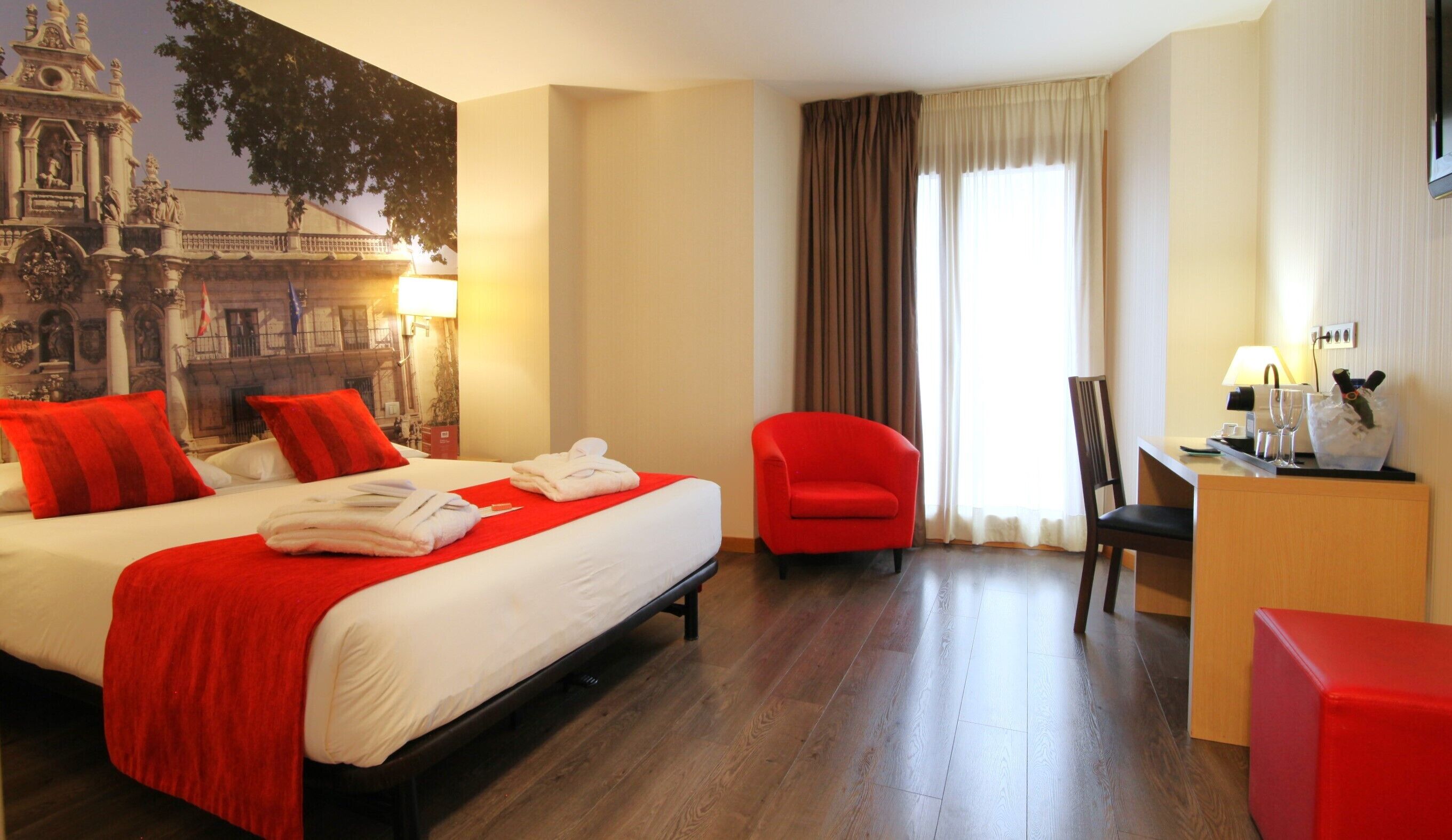 Habitación de hotel con una cama grande, almohadones rojos, un sillón rojo, un escritorio con lámpara y vistas a un edificio a través de la ventana.