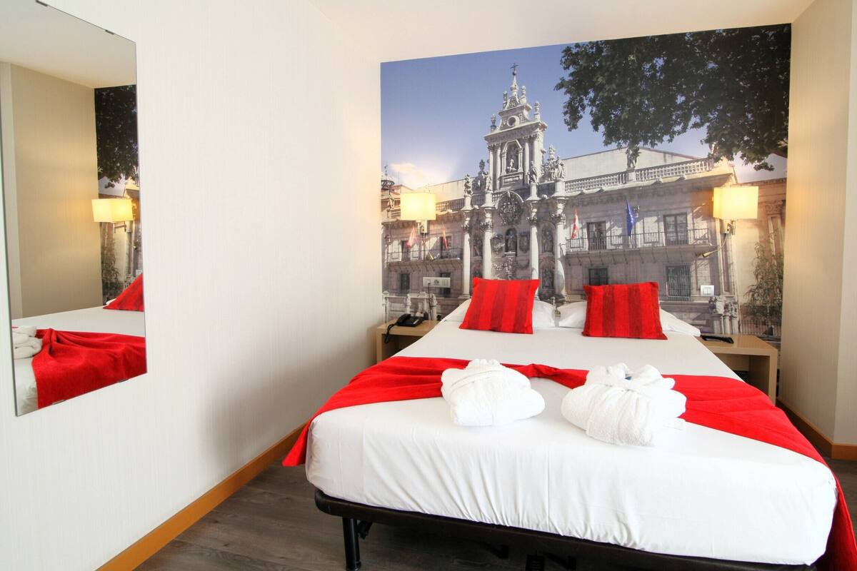 A hotel room with a large bed, red pillows, and a mural of a building.