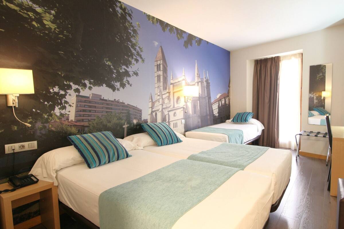 A hotel room with two beds, a desk, a chair, a TV, and a large mural of a cathedral.