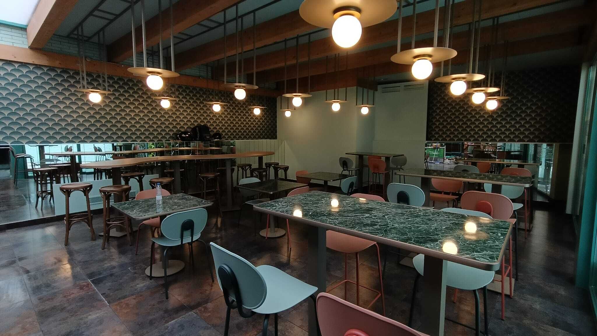 Interior moderno de restaurante con techos altos, vigas a la vista y una zona de bar central. Mesas y sillas distribuidas por todo el espacio, con un papel tapiz estampado en el fondo.
