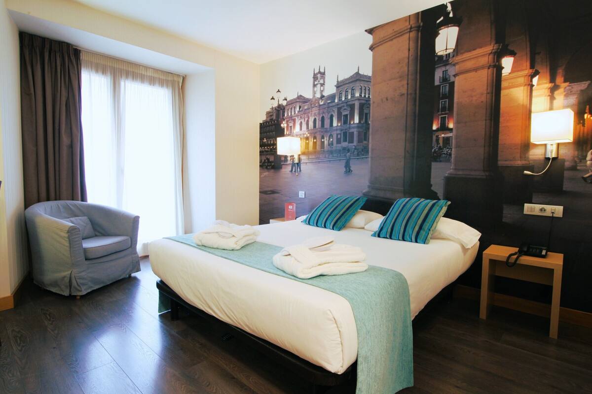 A hotel room with a large bed, a chair, a nightstand, and a wall mural of a cityscape.