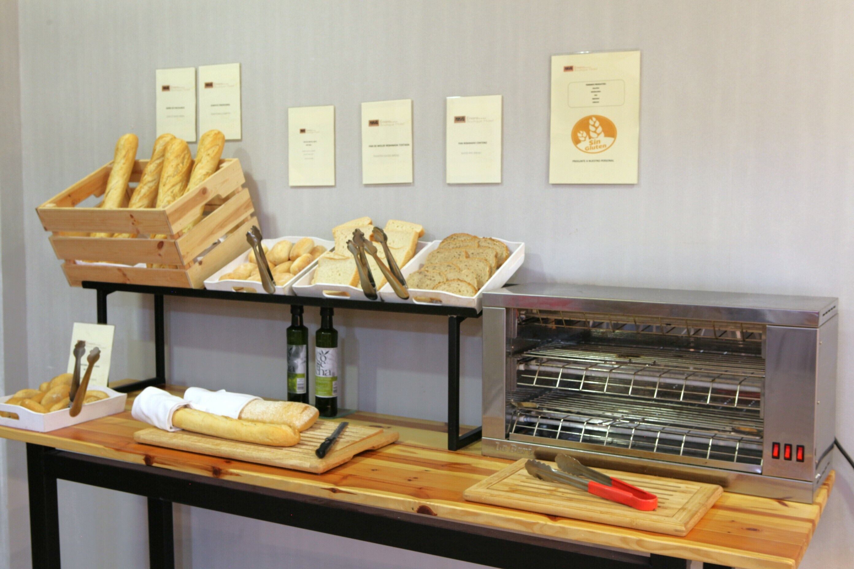 Una vitrina de panadería con pan, barras y bollos sobre una mesa de madera, con un horno de pan y diplomas en la pared.