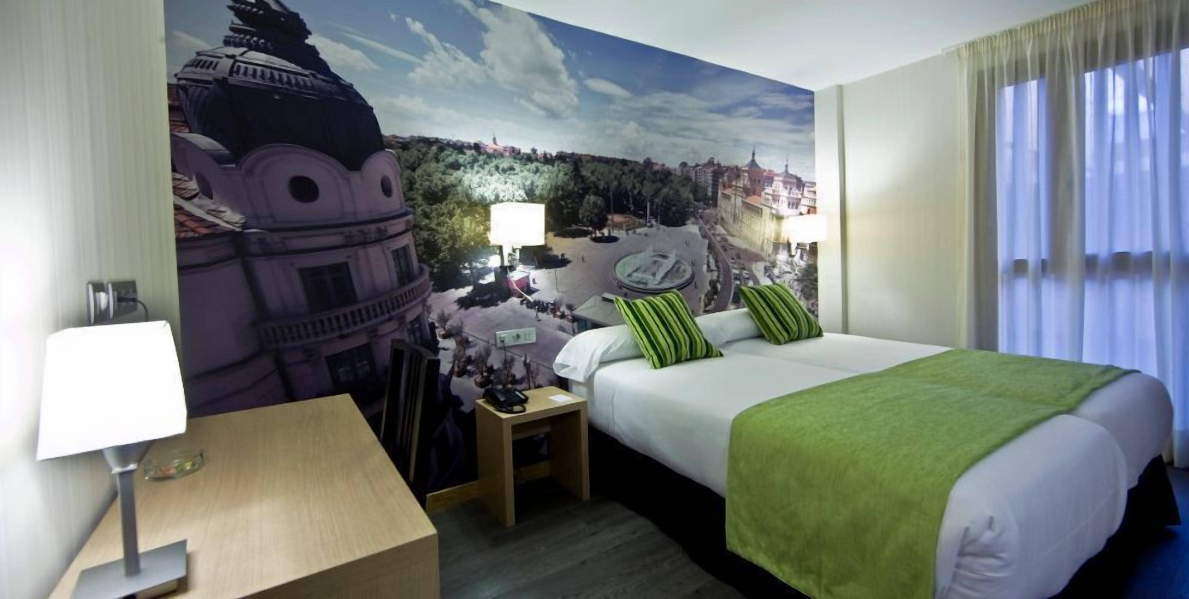 A hotel room with a large mural of a cityscape, a bed with green bedding, a bedside table with a lamp, and a window with curtains.