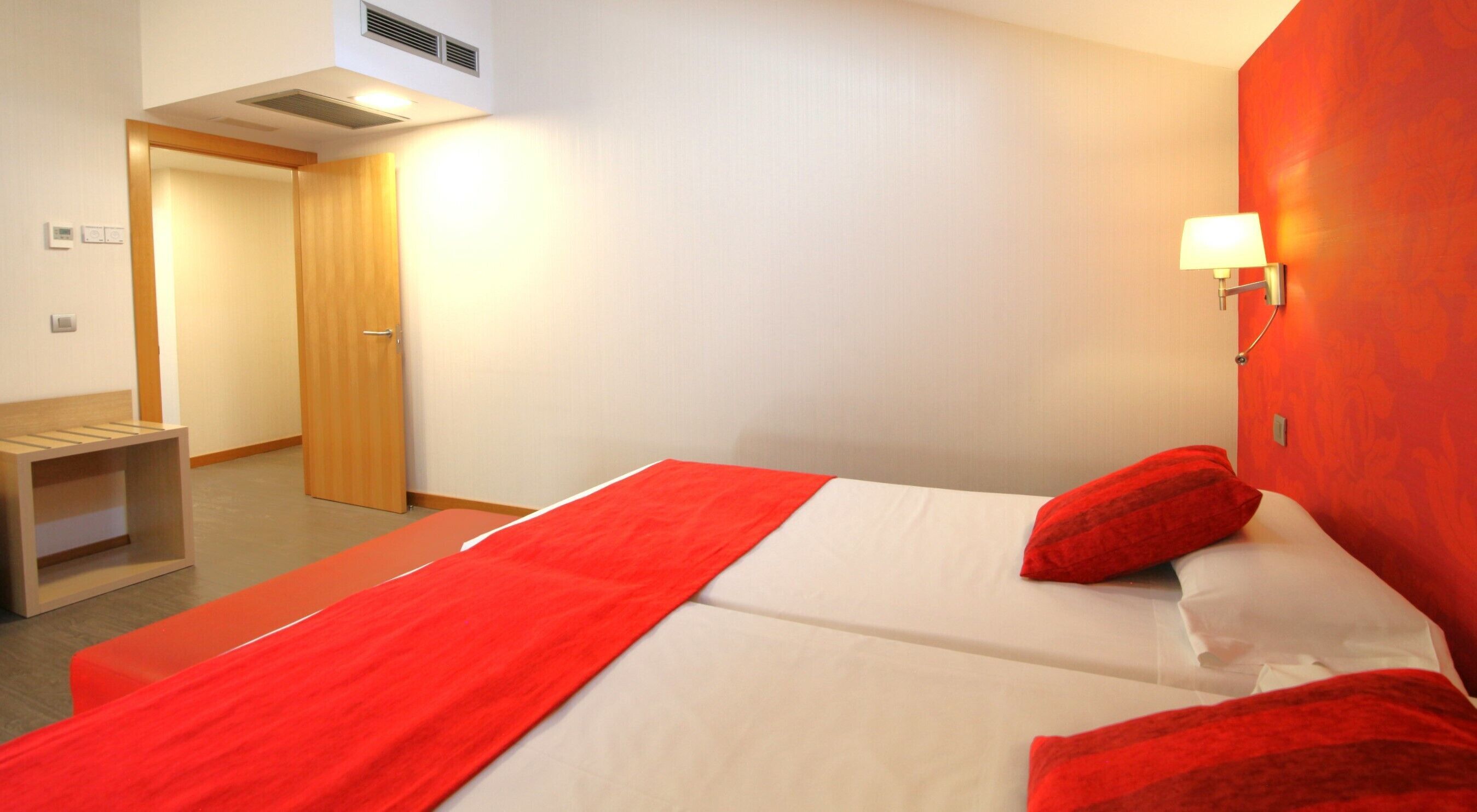 Una habitación de hotel con cama, una manta roja, almohada roja, mesita de noche y una lámpara fijada a la pared.