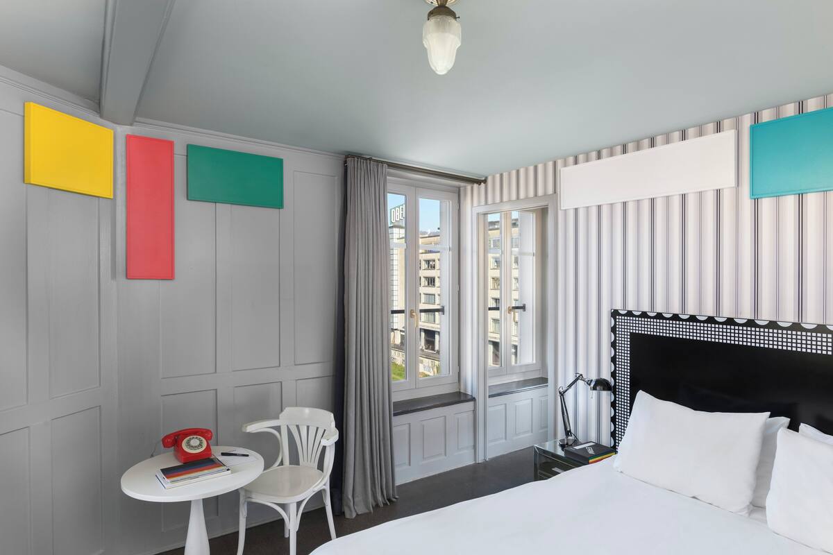 A hotel room with a bed, a small table with a telephone, a chair, and a window with curtains.