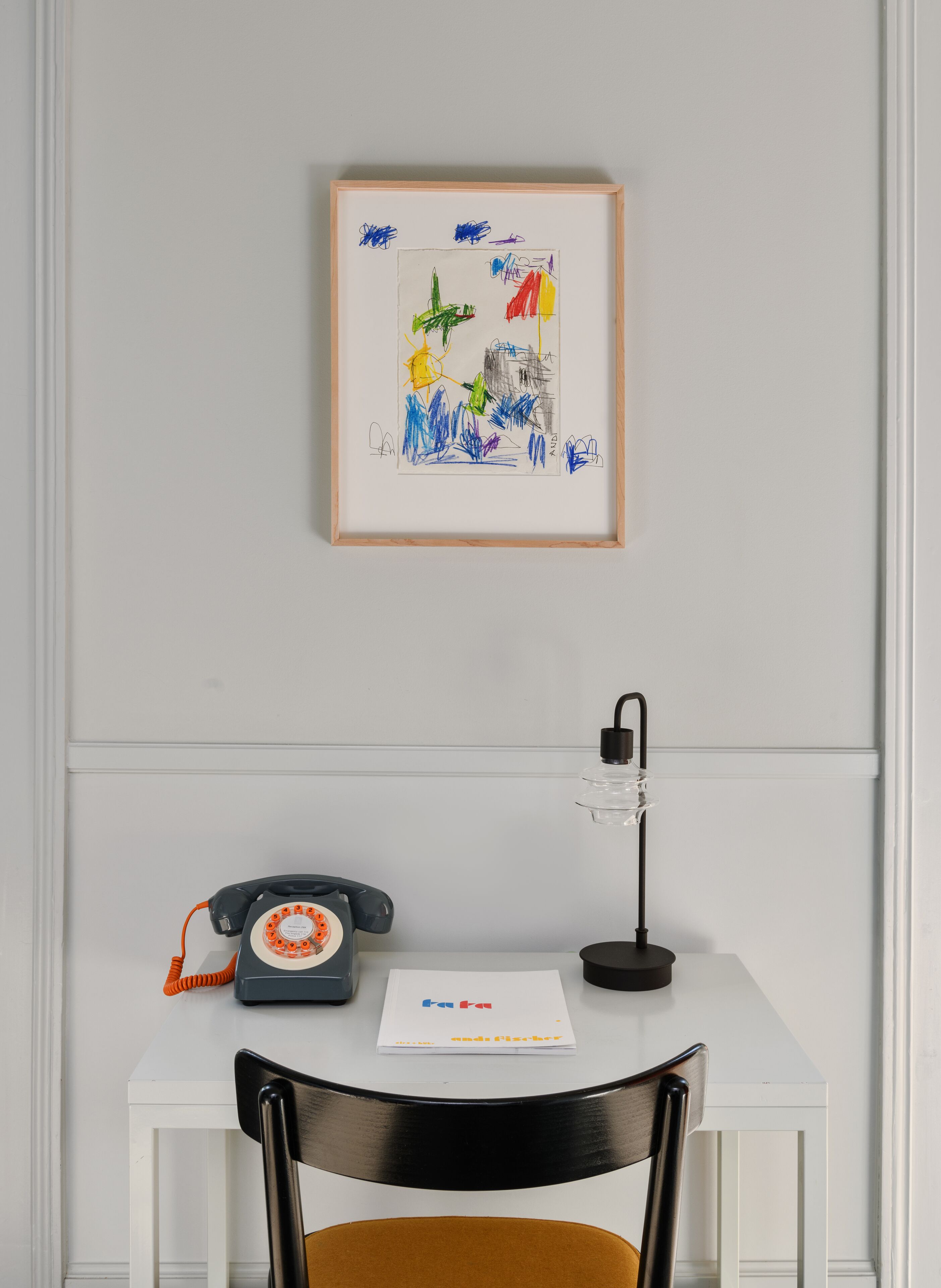Un bureau blanc avec un téléphone noir, une lampe de bureau noire et un dessin encadré accroché au mur.