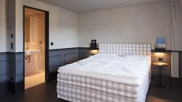 A hotel room with a large bed, a bedside table, a nightstand, a wall-mounted shelf, and a bathroom with a sink and mirror.
