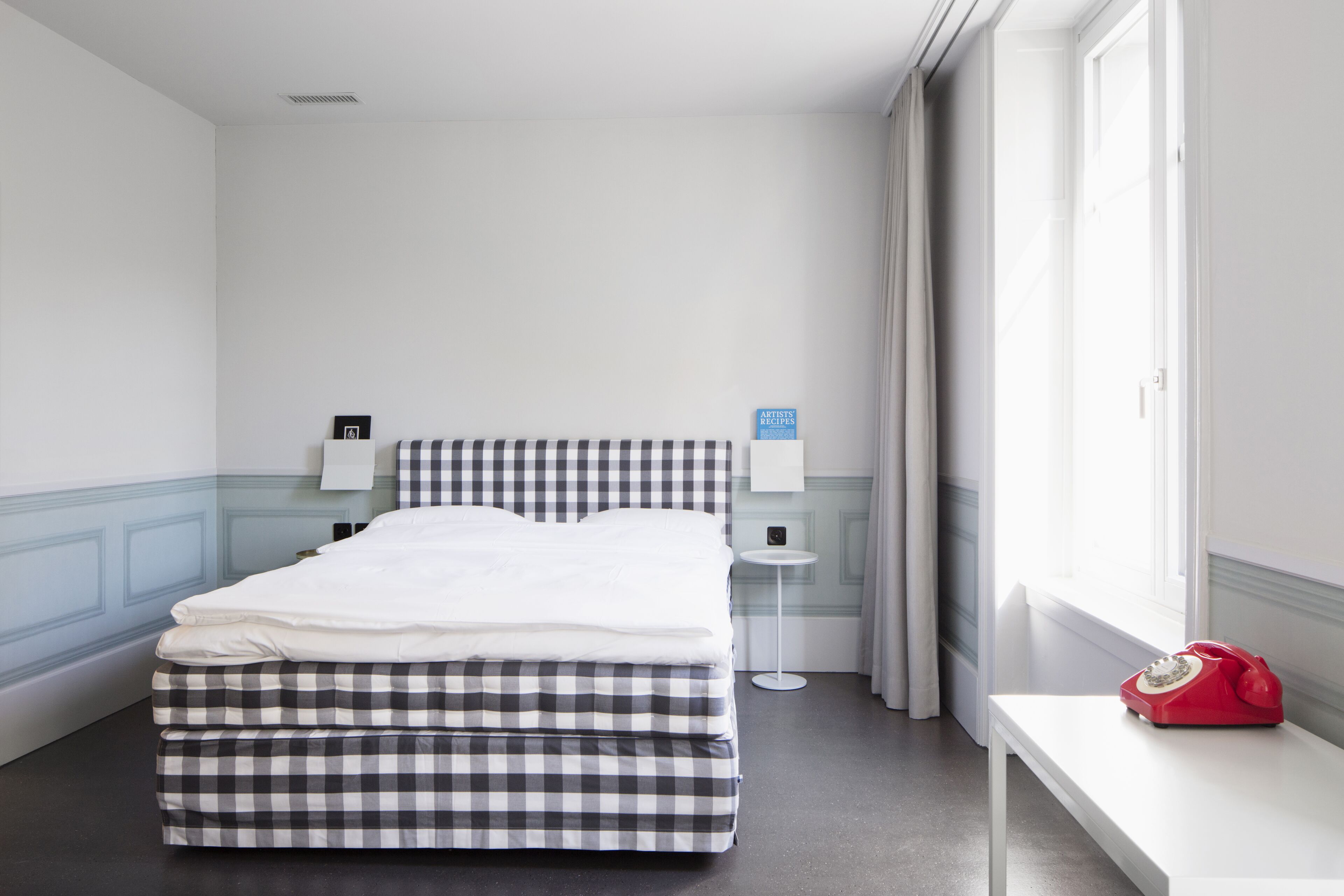 A hotel room with a checkered bed, a bedside table with a red telephone, and a window with curtains.
