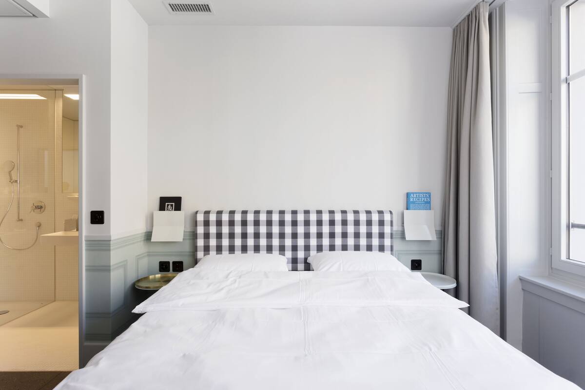A hotel room with a checkered headboard, a bed with white linens, a bedside table with a lamp, a small round table, and a window with curtains.