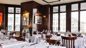 A restaurant with multiple tables set for dining, large windows offering a view of buildings, and a chandelier hanging from the ceiling. - Boutique & Art Hotel Helvetia (Zürich)
