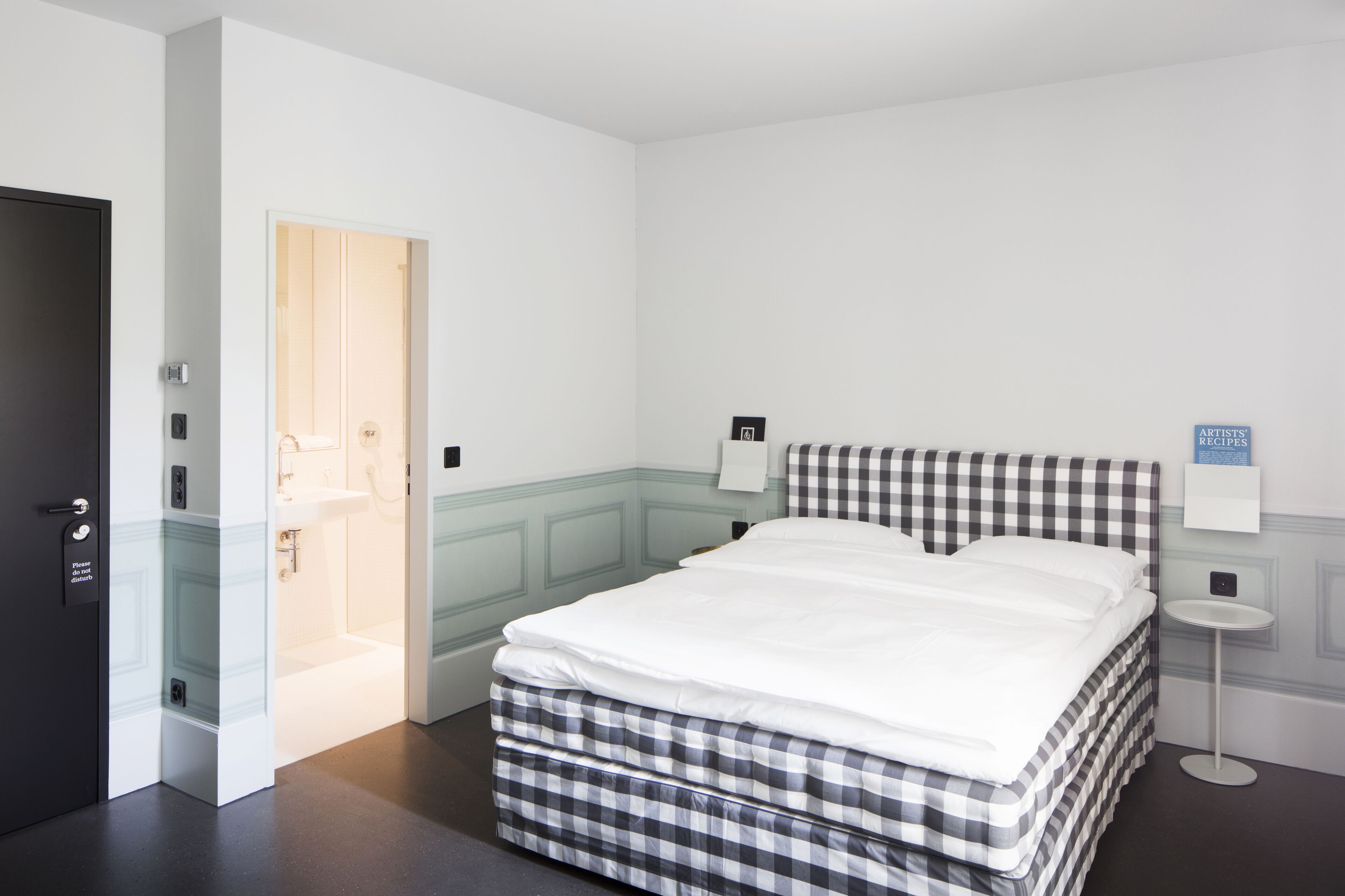 A neatly arranged hotel room with a bed, a small bedside table, a wall-mounted sign, and a door leading to a bathroom.