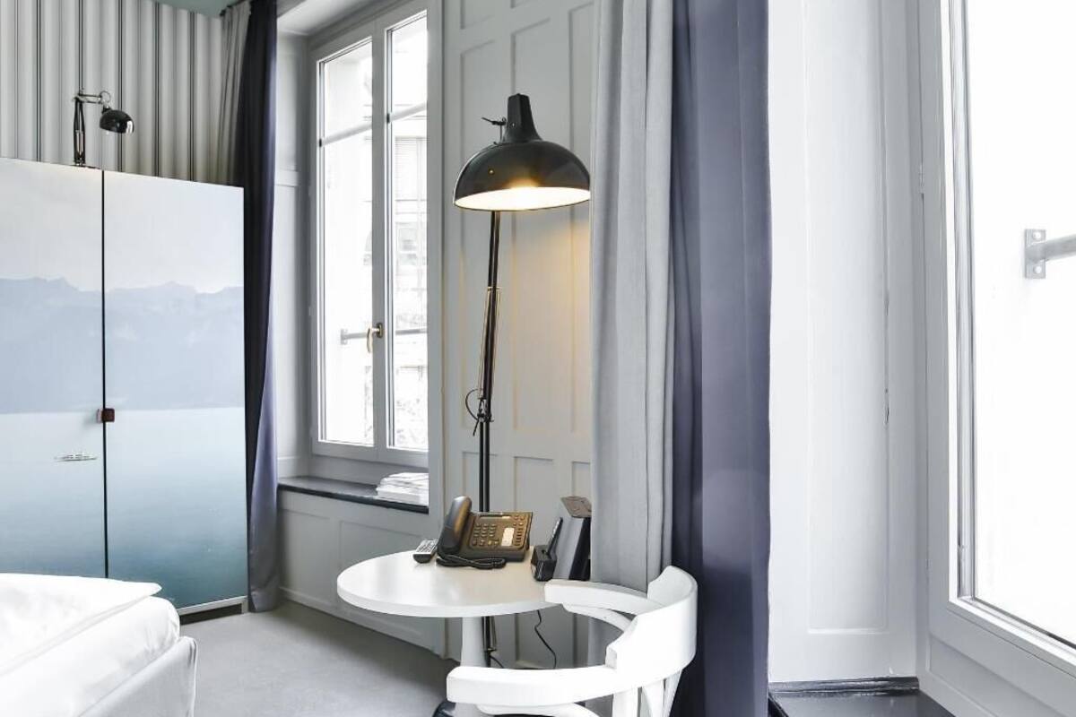 A hotel room with a bed, a desk with a telephone, a lamp, and a view through a window.