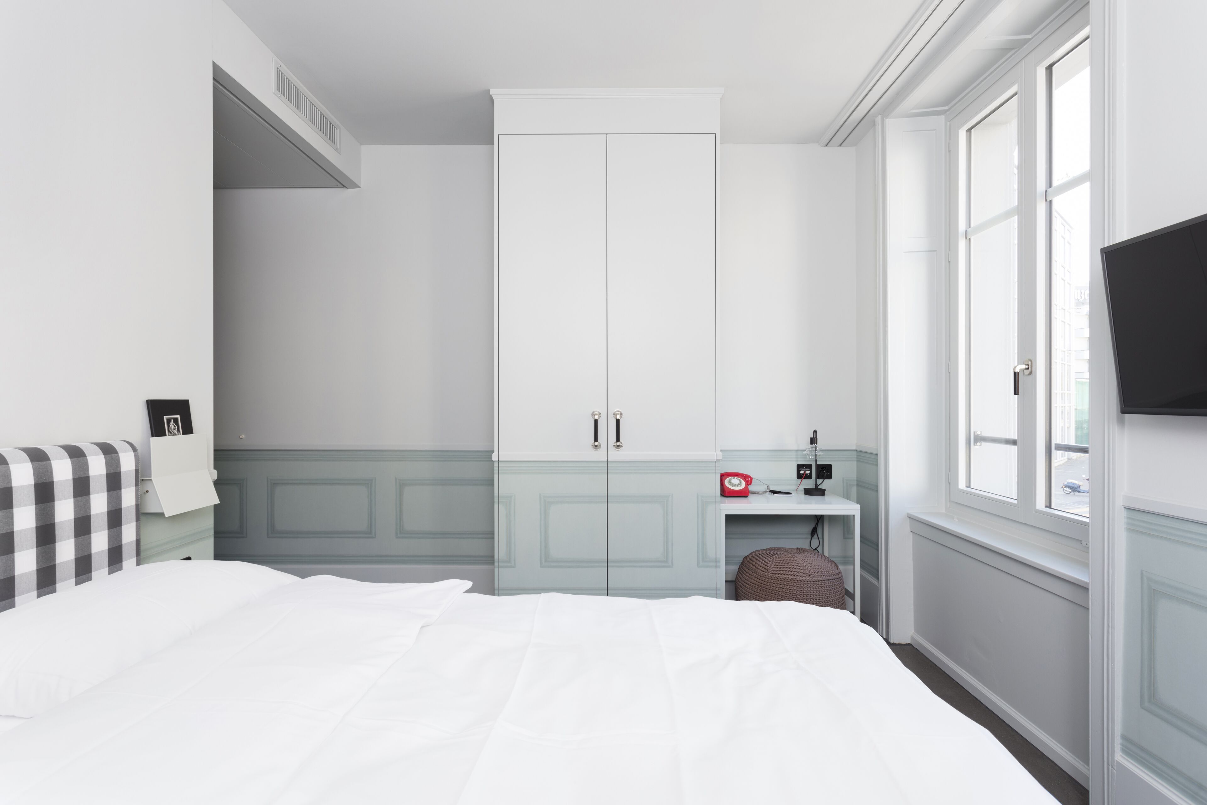A bedroom with a bed, a wardrobe, a small table with a clock, and a window.