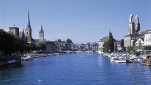 A river with boats, a bridge, and historic buildings including a church with a tall spire and a cathedral with two domes. - Boutique & Art Hotel Helvetia (Zürich)