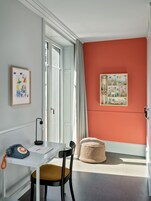 Um quarto com uma mesa branca, uma cadeira preta, um banquinho de vime e um quadro pendurado na parede.