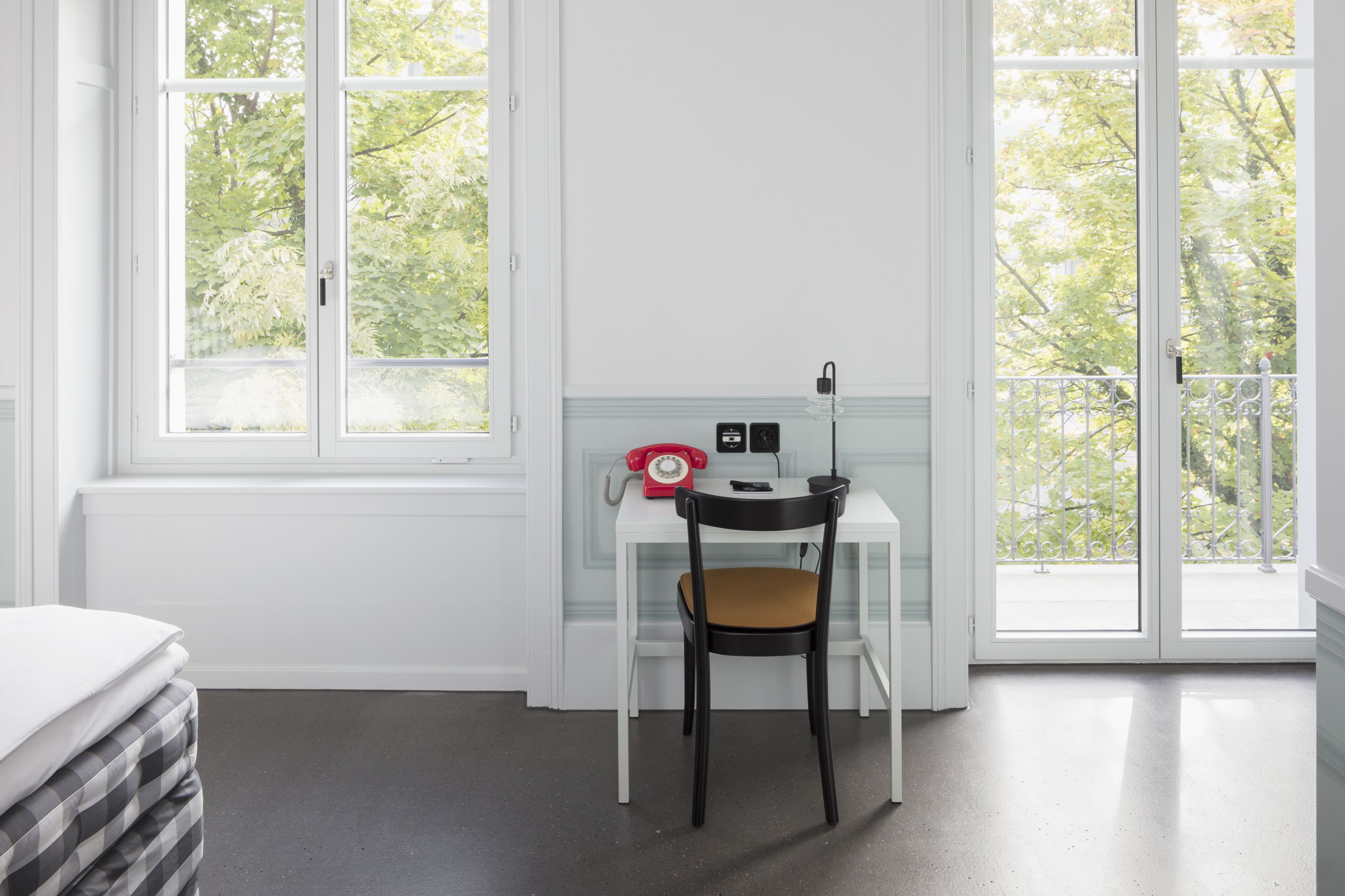 A room with a white desk, a black chair, a red rotary phone, and a window with a view of trees.