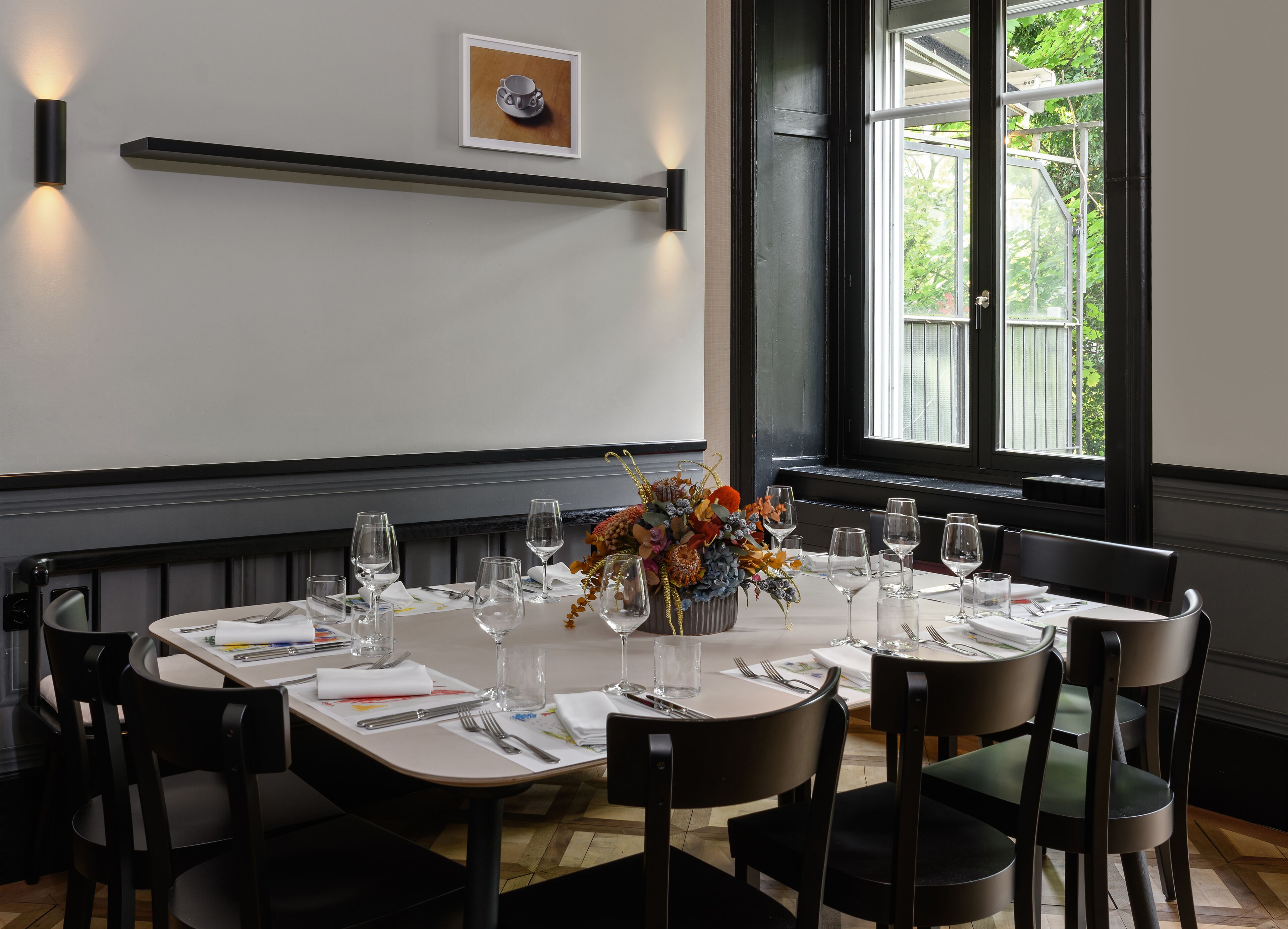 Un coin repas avec une table dressée pour six personnes, des chaises, un vase de fleurs et un tableau encadré accroché au mur.
